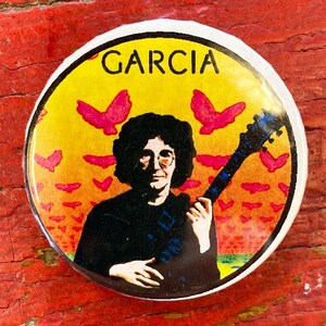 May include: Round button with the name "GARCIA" in black letters at the top. The button features an illustration of a person with glasses holding a guitar, set against a yellow and orange background with pink bird shapes.