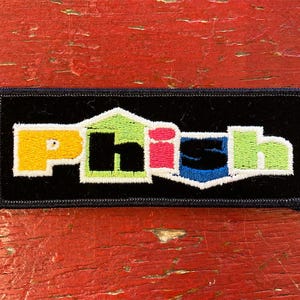 May include: A black embroidered patch featuring the word "Phish" in colourful letters. The letters are in a stylised font and outlined in white. The patch is rectangular and has a black border.