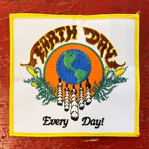 May include: A white embroidered patch with a yellow border. The patch features brown stylised text that reads "Earth Day" in a circle around a blue globe with green continents. The globe is surrounded by a circle of orange and green leaves. Below the globe are five white feathers. The text "Every Day!" is written in cursive below the feathers.