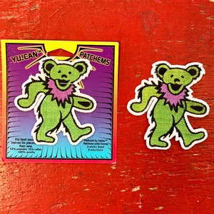 May include: Green and purple embroidered iron-on patch of a smiling bear with a pink collar. The patch is on a card with the text "YUJEAN PATCHEMS" and instructions for applying the patch. Produced by YuJean Patches under license Grateful Dead Productions.