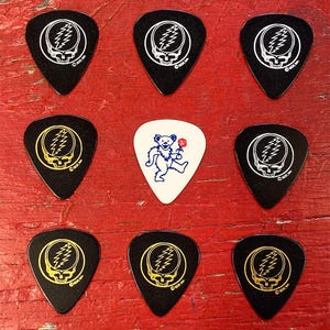 May include: Nine guitar picks with a Grateful Dead logo. Eight picks are black with a white or gold logo. One pick is white with a blue logo of a dancing bear holding a flower.