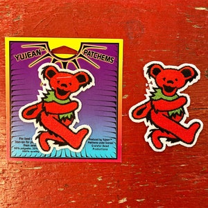May include: Red and green Grateful Dead dancing bear iron-on patch on a white background. The patch is on a card with the text "YUJEAN™ PATCHEMS" and "Produced by Yujean™ Palchems under license Grateful Dead Productions".
