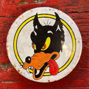 May include: A white round button with a yellow circle around a cartoon wolf with a red tongue sticking out. The wolf has black fur, yellow eyes, and an orange nose.
