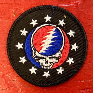 May include: A black circular patch with a white border featuring a circle of white stars around a red, white, and blue Grateful Dead Steal Your Face logo.