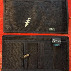 Könnte beinhalten: Schwarze Hanf-Geldbörse mit weißem Blitzdesign und dem Wort "HEMP". Die offene Geldbörse zeigt mehrere Fächer, darunter eine Reißverschlusstasche. Das Etikett lautet "Artisan Grateful Dead Product".