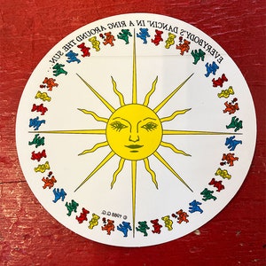 May include: A white circular sticker with a yellow sun in the center and a ring of dancing bears around the sun. The text "EVERYBODY'S DANCIN' IN A RING AROUND THE SUN." is written around the outside of the sticker.