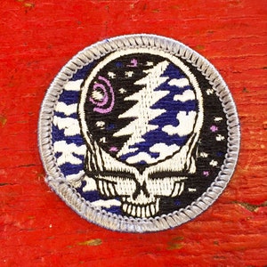 May include: A round, grey embroidered patch featuring a white and blue Grateful Dead Steal Your Face logo. The logo is surrounded by a black background with white stars and purple swirls.