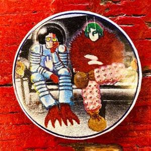May include: A round button featuring a colorful illustration of two figures. One figure is wearing a blue and white space suit with red claws. The other figure is wearing a red and pink outfit with a green head.