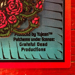 May include: A close-up of a red, black, and green embroidered rose patch. The patch has the text "Produced by YujeanTM Patches under license: Grateful Dead Productions".