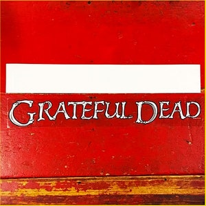 May include: A white sticker with the words "GRATEFUL DEAD" in a skeletal font on a red wooden surface.