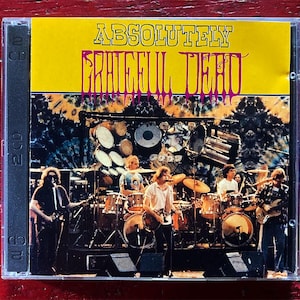 May include: A CD case featuring the album "Absolutely Dead" by the Grateful Dead. The cover has a yellow background with the band's name in pink and a tie-dye design. The image shows the band performing live.