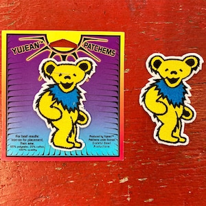 May include: Yellow smiling bear patch with a blue collar. The patch is on a purple and white background with the text "Yujean Patches" and "Produced by Yujean™ Palchems under license Grateful Dead Productions".