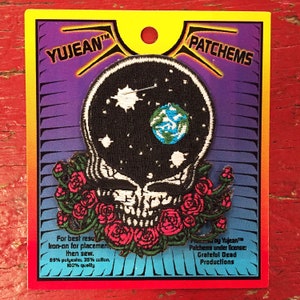 May include: A black and white embroidered patch of a skull with a galaxy inside and a small blue and green earth. The skull is surrounded by a wreath of red roses. The patch is on a card with the text "YUJEAN&trade; PATCHEMS" and "Proby Yujean&trade; Palchems under license: Grateful Dead Productions".