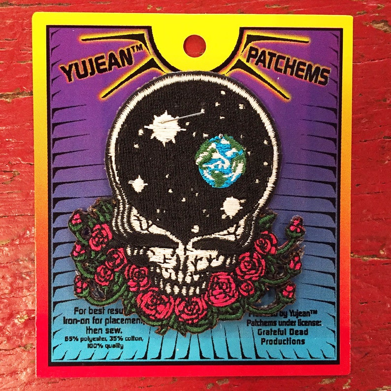 May include: A black and white embroidered patch of a skull with a galaxy inside and a small blue and green earth. The skull is surrounded by a wreath of red roses. The patch is on a card with the text "YUJEAN&trade; PATCHEMS" and "Proby Yujean&trade; Palchems under license: Grateful Dead Productions".