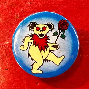 以下が含まれることがあります： 赤いサングラスと赤い羽根の襟をした黄色の漫画のクマが、赤いバラを持って歩いています。クマは青い背景の上にいます。