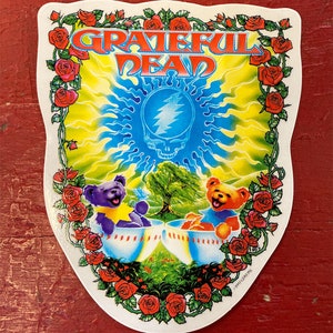 May include: A white sticker with a colorful Grateful Dead design. The sticker features a yellow sun with a blue lightning bolt, two dancing bears, and a wreath of red roses.