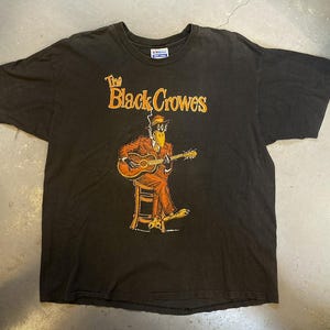Op de afbeelding: Zwart t-shirt met de tekst "The Black Crowes" in oranje. Daaronder een illustratie van een vogelachtig figuur in een oranje pak die gitaar speelt terwijl hij op een kruk zit.