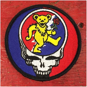 Peut inclure: Un écusson rond brodé représentant un ours jaune dansant tenant une rose rouge dans sa main, à l'intérieur d'un crâne blanc sur fond bleu et rouge.