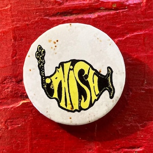 以下が含まれることがあります： 黄色と黒の魚のデザインが施された白いボタン。魚は「FISH」という言葉のようにスタイリッシュにデザインされており、尾が上向きにカールしています。