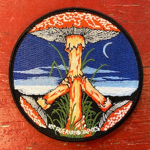 May include: A round embroidered patch featuring a peace sign formed by three red and white mushrooms, set against a blue sky with white clouds. The text "Not Fade Away Graphics" is at the bottom of the patch.