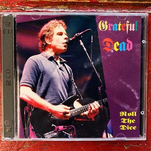 May include: A Grateful Dead CD case featuring a live performance image. The CD case has the band's name in colorful text at the top and the album title "Roll The Dice" in gold text at the bottom. The artist is playing a black guitar and singing into a microphone.