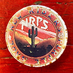 May include: A vintage, round, metal pin with a red, orange, and yellow sunset scene. The pin features a cactus and the text "NEW RIDERS OF THE PURPLE SAGE" and "NRPS".