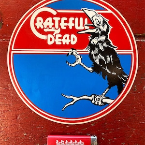 May include: A round sticker with a red and blue design featuring the words "Grateful Dead" and a black crow illustration. A red lighter with a white graphic of dancing figures is also present.