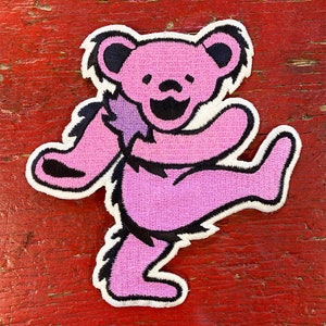May include: Pink dancing bear patch with black outline and a white border. The bear is wearing a purple bandana and has a big smile.
