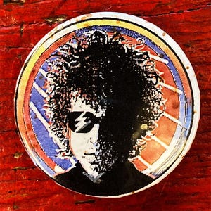 May include: A round button with a colourful design featuring a black and white image of a person with curly hair wearing sunglasses. The background is a red, white and blue design.