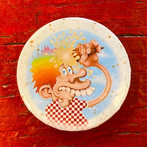 May include: A round, white button featuring a cartoon image of a person with rainbow hair being hit in the face with a cream pie. The person is wearing a red and white checked shirt.