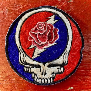 May include: A circular embroidered patch featuring a central red rose layered over a white lightning bolt. The design is set against a blue background, framed by a white border and a stylized white skull shape at the bottom, all outlined in black.