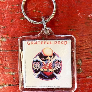 Może przedstawiać: Przezroczysty plastikowy brelok do kluczy z logo Grateful Dead: czaszka z czerwonym sercem, różami i skrzyżowanymi kośćmi. Brelok ma srebrny metalowy pierścień. Tło to czerwona, zwietrzała drewniana powierzchnia.