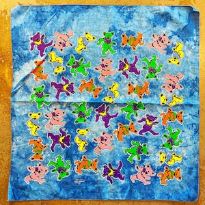 May include: A blue bandana with a tie-dye pattern featuring a repeating design of dancing bears in various colors. The bears are wearing hats and have smiling faces.