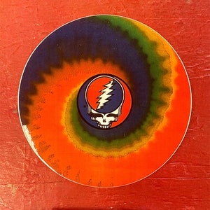 May include: A round sticker with a tie-dye pattern in shades of orange, yellow, green, and blue. The center of the sticker features a black and white skull with a lightning bolt design.