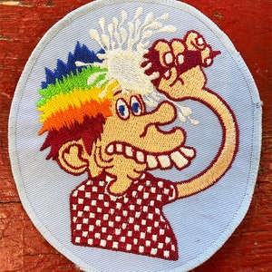 May include: A blue embroidered patch featuring a cartoon character with rainbow hair, a chequered shirt, and a white substance splattered on their head. The character is holding a bottle of white substance in their right hand.
