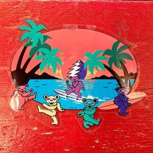 Puede incluir: Una pegatina con cinco osos de dibujos animados que llevan gafas de sol y montan tablas de surf. Los osos son de varios colores, incluyendo rosa, verde, azul y morado. El fondo es una escena de puesta de sol con palmeras y un océano azul.