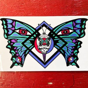 May include: A colourful sticker featuring a butterfly with blue, green and purple wings. The butterfly's eyes are red, and it is centred on a blue diamond shape. The butterfly's wings are spread wide, and the diamond shape is outlined in black.