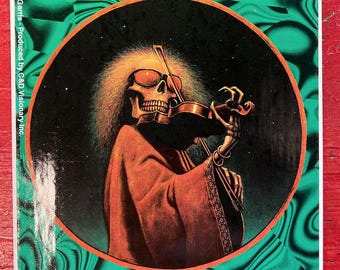 May include: A rectangular sticker featuring a circular illustration of a skeleton with frizzy hair and sunglasses playing a violin. The skeleton wears a dark robe. The background is a black starry sky, framed by a green and black wavy pattern. Text reads: 1975 Philip Garris - Produced by C&D Visionary Inc.