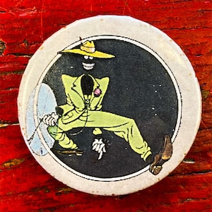 May include: A round button with a white border and a black background. A cartoon figure in a yellow hat and green suit is dancing. The figure's face is black with a white smile.