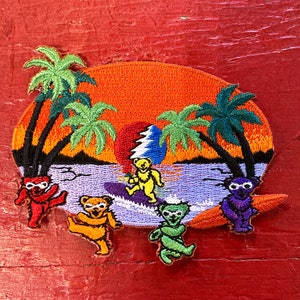 May include: Embroidered patch depicting a sunset scene with palm trees and four colourful bears riding waves on surfboards. The bears are wearing different coloured hats and have a psychedelic swirl design on their bellies.