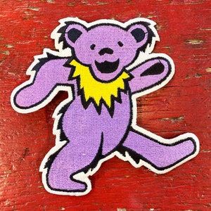 May include: Purple and yellow embroidered patch of a smiling bear with a fuzzy outline. The bear is walking with its right leg forward and its left arm raised.