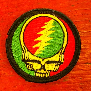 Peut inclure: Un patch rond brodé avec un contour noir. Le centre présente un tourbillon vert et rouge avec un éclair jaune. La moitié inférieure du patch montre un crâne jaune avec des yeux et des dents noirs.