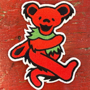 May include: Red and green embroidered patch of a smiling bear wearing a green scarf. The bear is standing on its hind legs with its arms raised.
