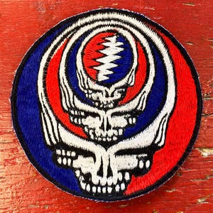 May include: A circular embroidered patch with a red, white and blue design. The design features a stylised skull with a lightning bolt in the centre. The skull is surrounded by concentric circles of red, white and blue.