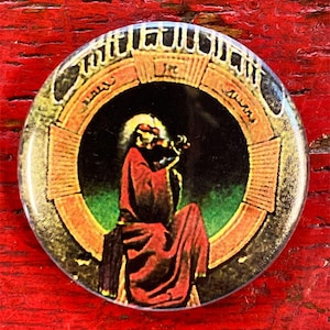 May include: A round button featuring a colourful image of a person in a red robe playing a violin. The image is surrounded by a circular frame with the words "In" and "Aeterno" written in gold.
