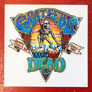 Könnte beinhalten: Ein bunter Grateful Dead-Aufkleber mit einem Skelett, das Schlagzeug spielt. Der Aufkleber zeigt die Worte "Grateful Dead" in einer stilisierten Schrift und den Text "Quality Rock & Roll" und "Since 1965" auf Bannern. Der Aufkleber enthält auch eine Bergkette und Rosen.
