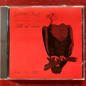May include: A CD case for the Grateful Dead's 'Little Red Rooster' album, featuring a red cover with a bald eagle illustration. Text includes 'Grateful Dead' and 'Live USA 1988'. The CD is housed in a clear plastic case.