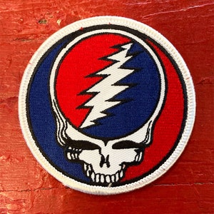 May include: A white, blue and red embroidered patch featuring a skull and lightning bolt design. The skull has a white face with black eyes and a black mouth. The lightning bolt is white and is in the centre of the patch. The patch is circular and has a white border.