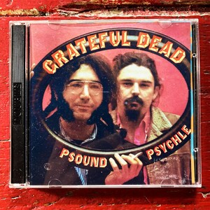 May include: A CD case featuring the band Grateful Dead. The album cover has a pink background with the band's name in orange lettering. Two men are in the center of the cover, inside of a black oval. The words "Psound Psychle" are also on the cover.