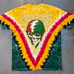 Könnte beinhalten: Gelbes Batik-T-Shirt mit Totenkopf-Grafik. Der Totenkopf hat einen rot-weiß-grünen Hut und Dreadlocks. Das Shirt hat ein V-förmiges Muster in Rot, Gelb und Grün.